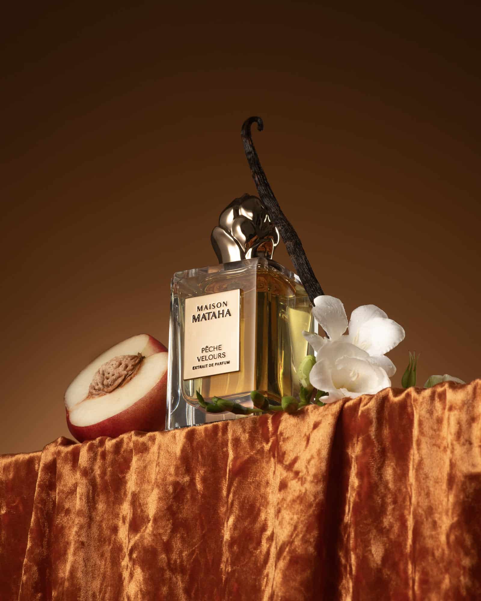 Flacon du parfum Pêche Velours de Maison Mataha posé sur un tissu orangé avec une pêche mûre, gousse de vanille et fleurs blanches, illustrant l’univers chaleureux et raffiné du parfum.