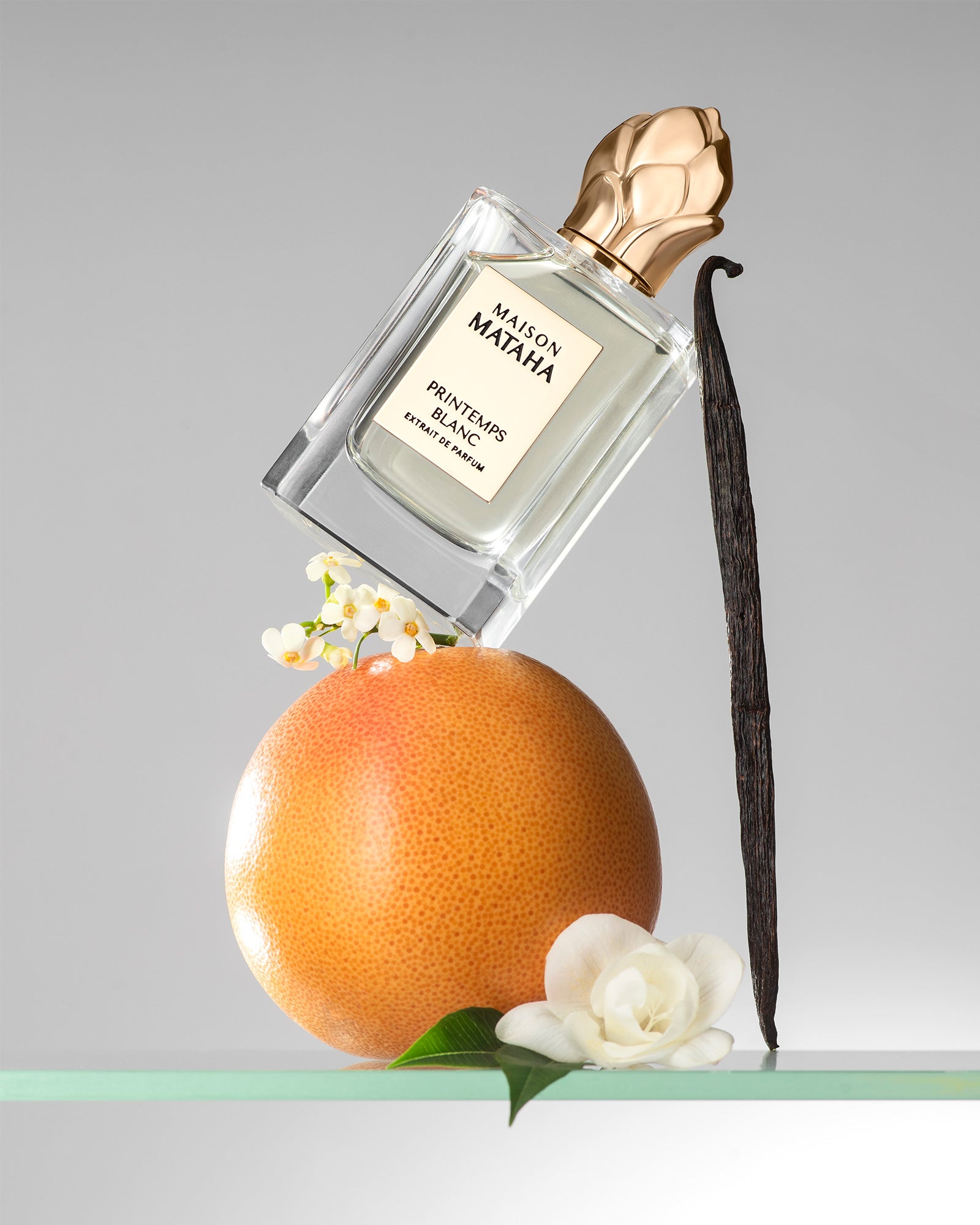 Flacon du parfum Printemps Blanc de Maison Mataha posé sur un pamplemousse, accompagné de fleurs blanches et d’une gousse de vanille