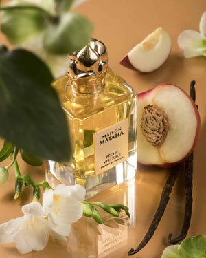 Bouteille de Pêche Velours par Maison Mataha posée sur fond doré avec fleurs blanches, pêche coupée et vanille, évoquant la gourmandise fruitée et la sensualité du parfum unisexe.